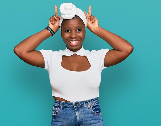 Young african woman with turban wearing hair turban over isolated background posing funny and crazy with fingers on head as bunny ears, smiling cheerful