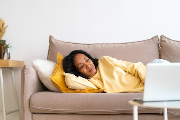 Young africanamerican female watching movies on laptop while lying on sofa in living room