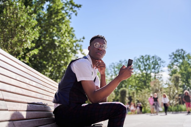 Young africanamerican man holding a phone and reading a message he is wearing glasses and sitting in an outdoor park