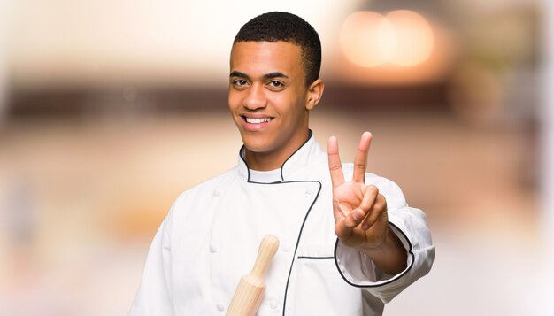 Young afro american chef man smiling and showing victory sign on unfocused background