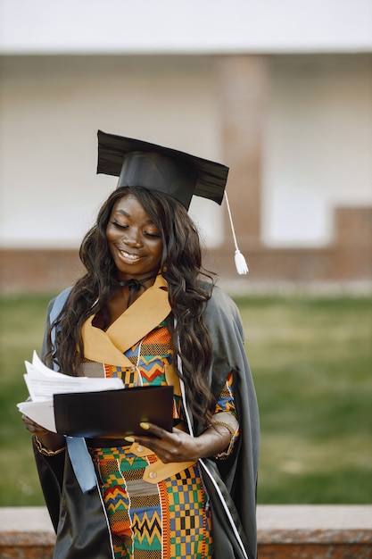 Young afro american female student dressed in black graduation gown. Campus as a background