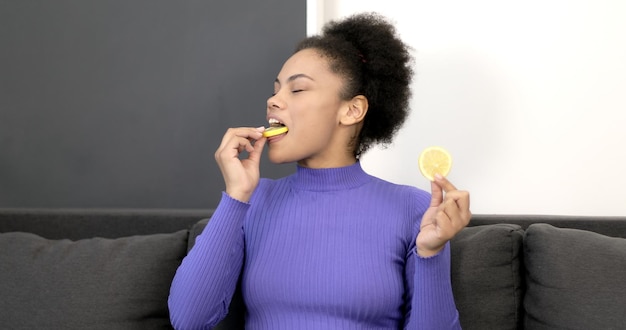 Young Afro american girl eating lemon from a plate on a sofa. Sour fruit. Healthy eating.