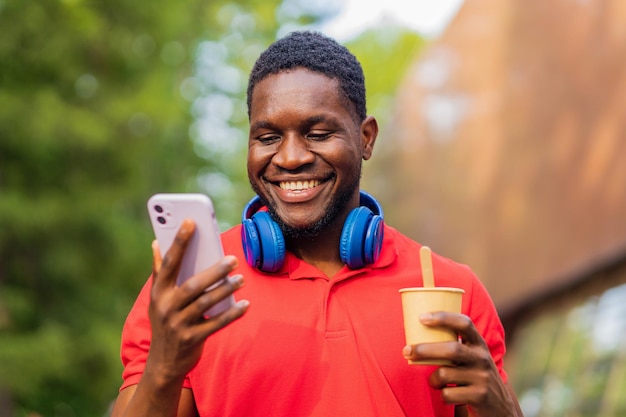 夏の公園でスマートフォンを使用して首にヘッドフォンを持つ若いアフロアメリカ人男性