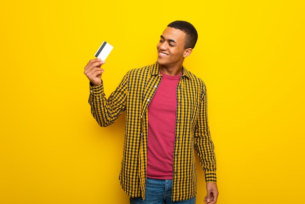 Young afro american man on yellow background holding a credit card and thinking