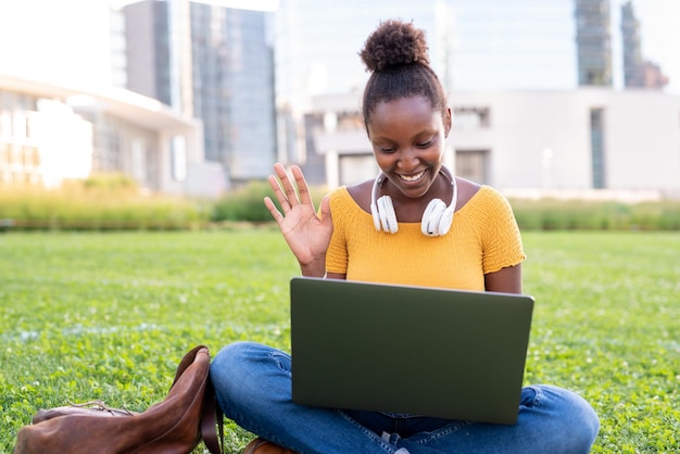 Young afro american student sitting on the lawn of the city park, woman engaged in outdoor video calling with her laptop