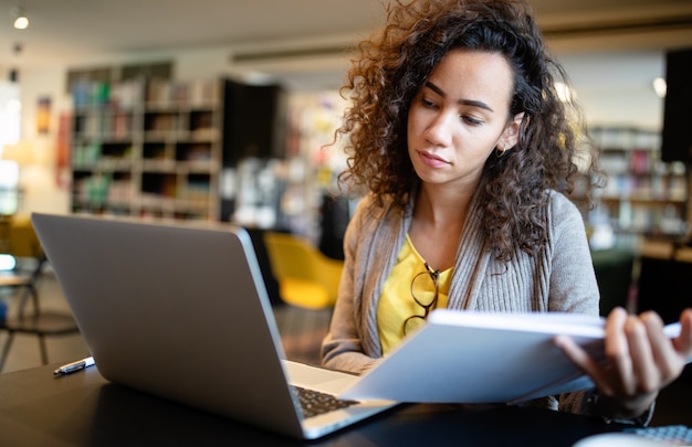 Giovane donna afroamericana seduta a tavola con libri e laptop per trovare informazioni. giovane studentessa che prende appunti da laptop e libri per il suo studio in biblioteca.