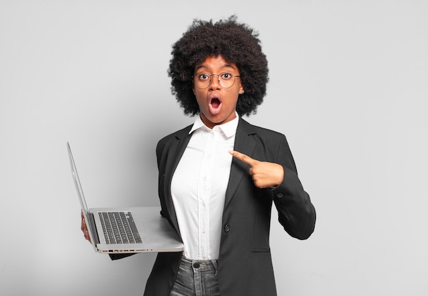 Young afro businesswoman looking shocked and surprised with mouth wide open, pointing to self