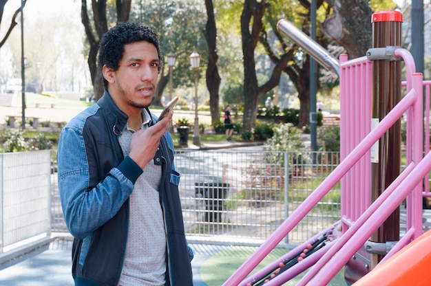 young afro latin man standing in public park sending voice note on his phone looking at camera