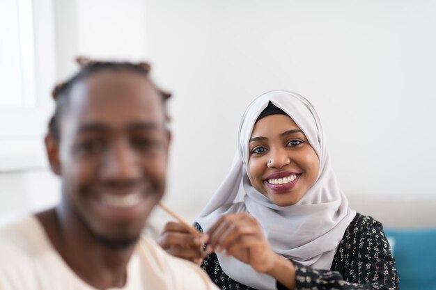 Photo young afro muslim couple have romantic time at home while woman making hairstyle to husband female wearing  traditional sudan islamic hijab clothes