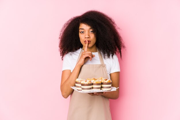 Young afro pastry maker woman holding a cupcakes isolatedYoung afro baker woman keeping a secret or asking for silence.