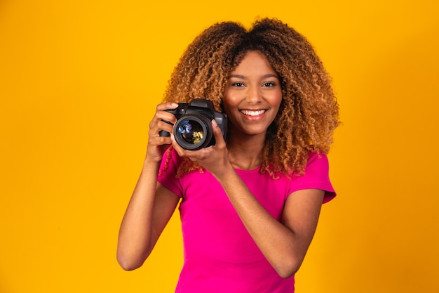 黄色の背景に手でカメラを保持している若いアフロ写真家。