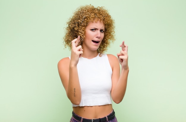 Giovane donna afro incrociando le dita con ansia e sperando in buona fortuna con uno sguardo preoccupato contro il muro verde