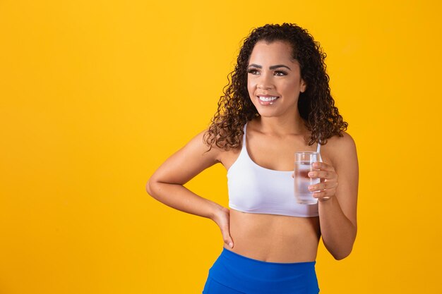 Young afro woman drinking water in yellow background. Young girl with glass of water. Young woman drinking water from the glass. Hydration concept