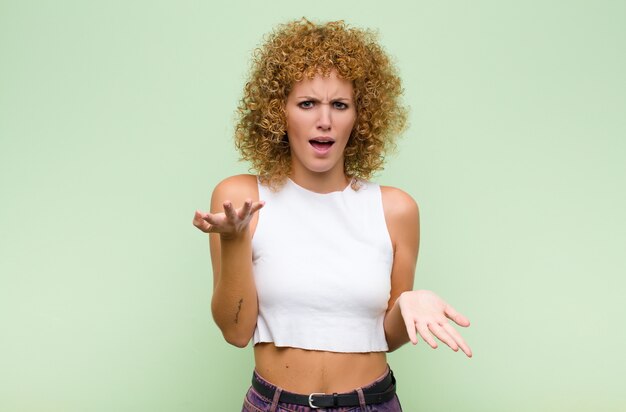 Young afro woman feeling clueless and confused, not sure which choice or option to pick, wondering against green wall