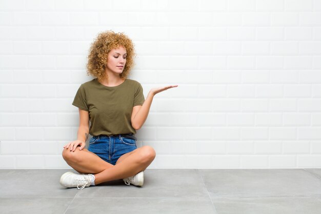 Young afro woman feeling happy and smiling casually, looking to an object or concept held on the hand on the side sitting on a floor