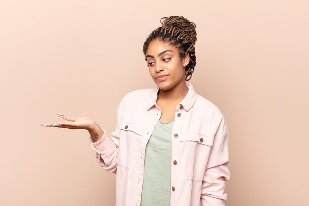 Young afro woman feeling happy and smiling casually, looking to an object or concept held on the hand on the side