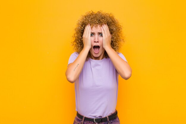 Young afro woman feeling horrified and shocked, raising hands to head and panicking at a mistake against orange wall