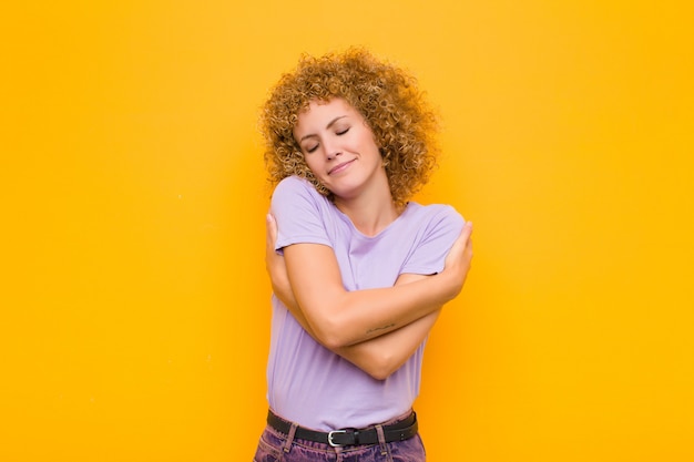 Giovane donna afro sentirsi innamorati, sorridente, coccole e abbracciarsi, restare single, essere egoisti ed egocentrici contro la parete arancione