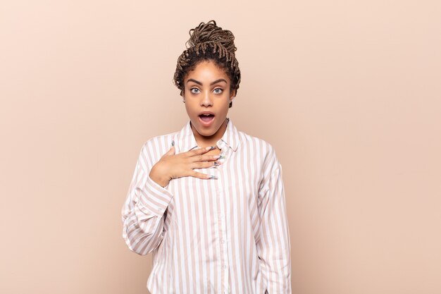 Young afro woman feeling shocked, astonished and surprised, with hand on chest and open mouth, saying who, me?