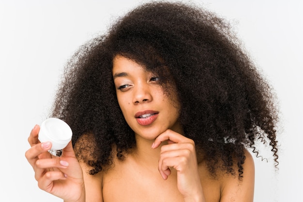 Young afro woman holding a moisturizer isolated looking sideways with doubtful and skeptical expression.