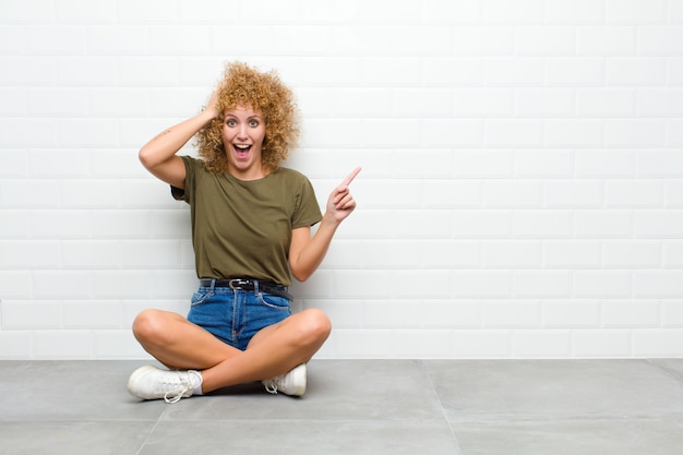 Giovane donna afro che ride, che sembra felice, positiva e sorpresa, realizzando una grande idea che punta allo spazio laterale della copia seduto su un pavimento