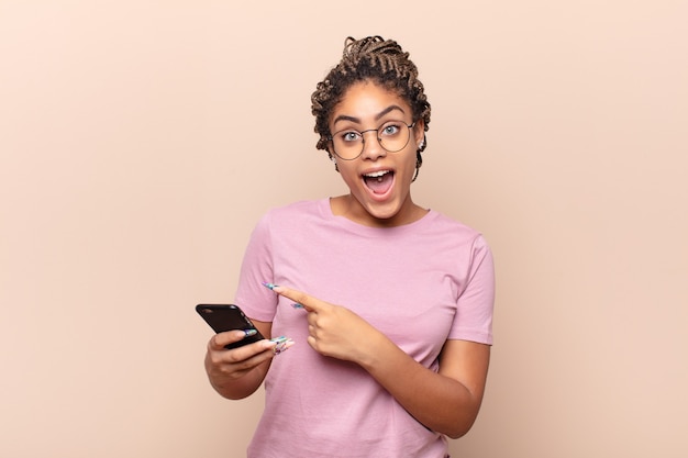 Young afro woman looking excited and surprised pointing to the side and upwards to copy space. smart phone concept