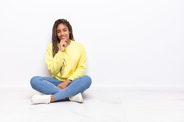 Young afro woman looking happy and smiling with hand on chin, wondering or asking a question, comparing options