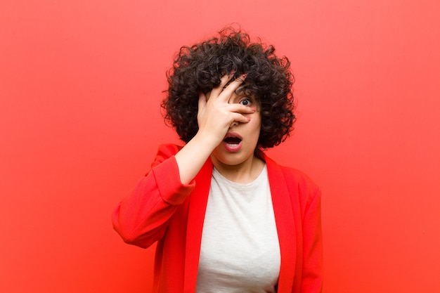 Young afro woman looking shocked, scared or terrified, covering face with hand and peeking between fingers