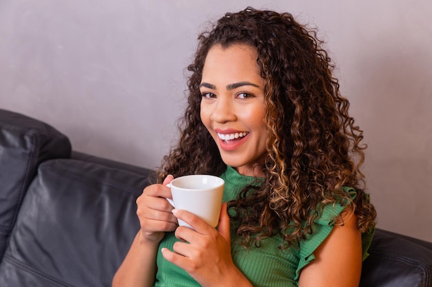 Young afro woman relaxing on the sofa having tea or coffee from
a cup.