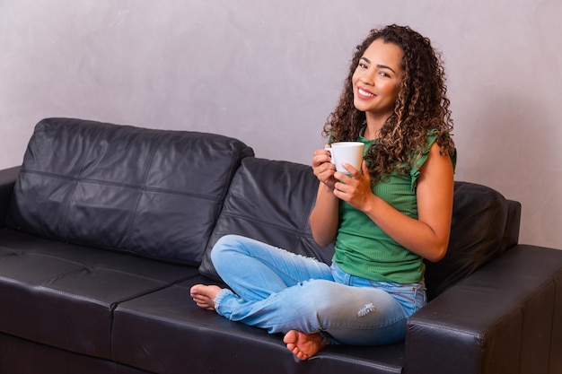 Young afro woman relaxing on the sofa having tea or coffee from
a cup.