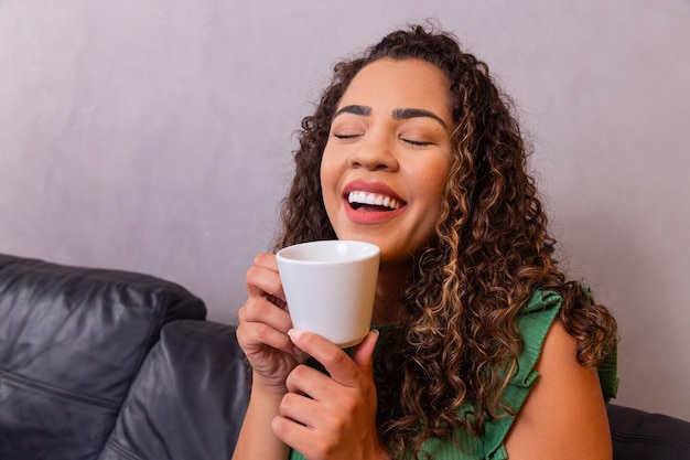 Young afro woman relaxing on the sofa having tea or coffee from\
a cup.