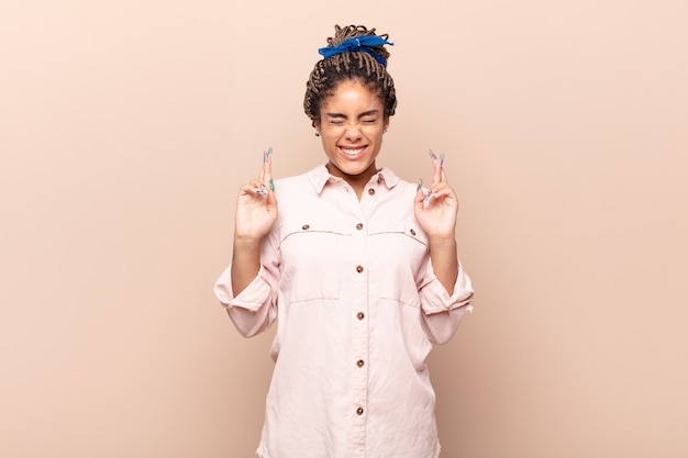 Young afro woman smiling and anxiously crossing both fingers, feeling worried and wishing or hoping for good luck