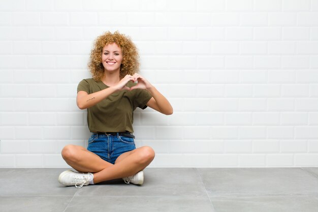 Photo young afro woman smiling and feeling happy, cute, romantic and in love, making heart shape with both hands sitting on a floor