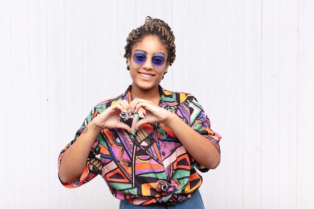 Young afro woman smiling and feeling happy, cute, romantic and in love, making heart shape with both hands