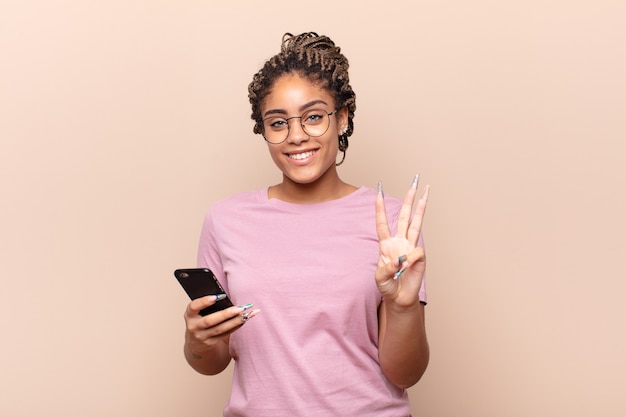 Giovane donna afro sorridente e guardando amichevole, mostrando il numero tre o terzo con la mano in avanti, il conto alla rovescia. concetto di smart phone