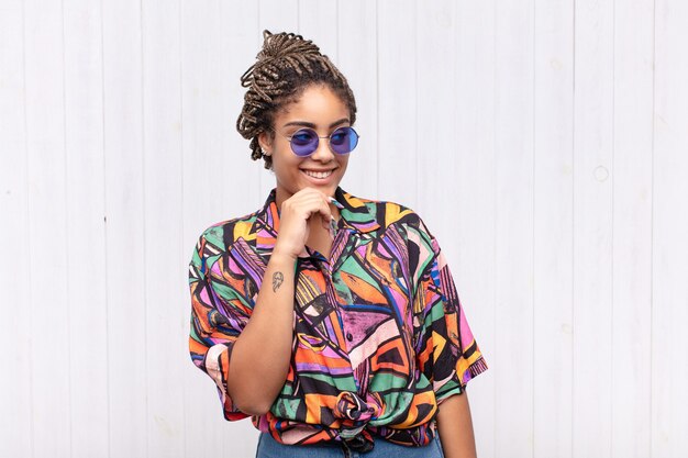 Young afro woman smiling with a happy, confident expression with hand on chin, wondering and looking to the side