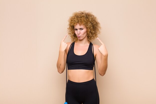 Young afro woman with a bad attitude looking proud and aggressive, pointing upwards or making fun sign with hands sport concept