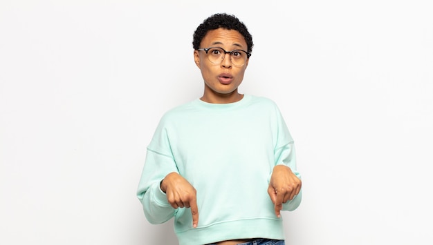 Young afro woman with open mouth pointing downwards with both hands, looking shocked, amazed and surprised