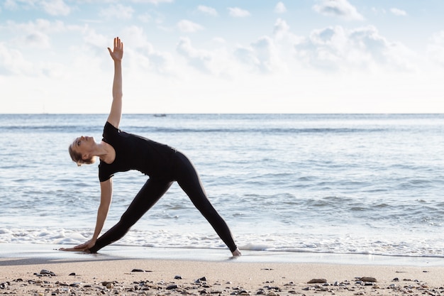 Young age woman in black doing yoga on sand beach