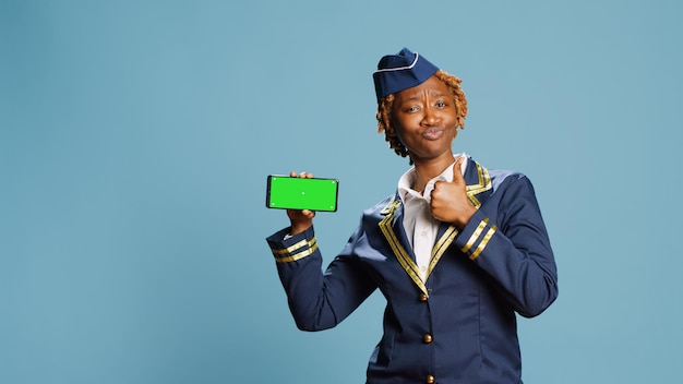Молодая стюардесса держит смартфон с зеленым экраном, показывая на камеру дисплей хроматического ключа с изолированным макетом. Женский член экипажа смотрит на пустой шаблон на синем фоне.