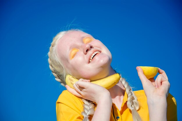 A young albino woman holds a banana to her ear like a phone against the blue sky