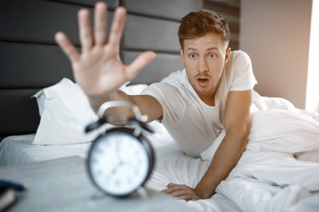 Young amazed man lyin on bed in morning. He overslept. Guy reach clock.