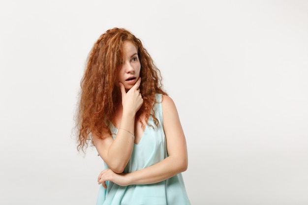 Giovane donna stupita rossa ragazza in abiti casual leggeri in posa isolata su sfondo bianco, ritratto in studio. concetto di stile di vita della gente. mock up copia spazio. metti la mano sul mento, guardando da parte.