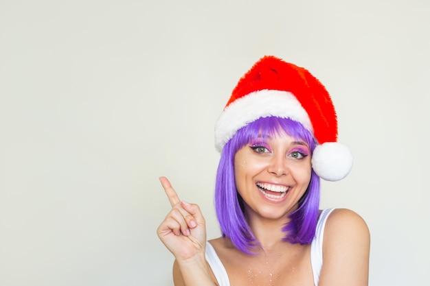 空のコピー スペースを指している赤いサンタ クロースの帽子をかぶった紫色の髪を持つ若い驚いた笑顔の女性