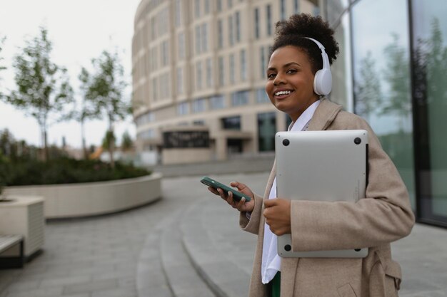 A young american woman with a wide smile walks in headphones with a mobile phone and a laptop