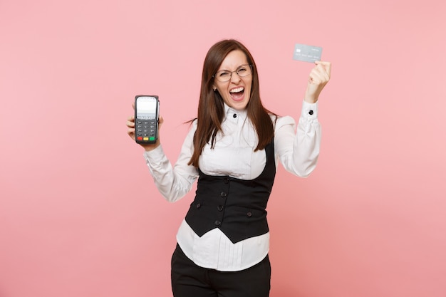 Young angry business woman scream hold wireless modern bank payment terminal to process and acquire credit card payments, black card isolated on pink background. Lady boss. Achievement career wealth.