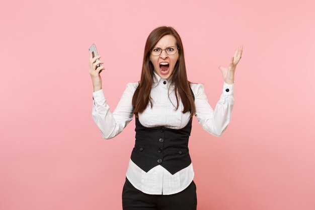 Young angry irritated business woman in glasses screaming holding mobile phone spreading hands isolated on pink background. Lady boss. Achievement career wealth concept. Copy space for advertisement.