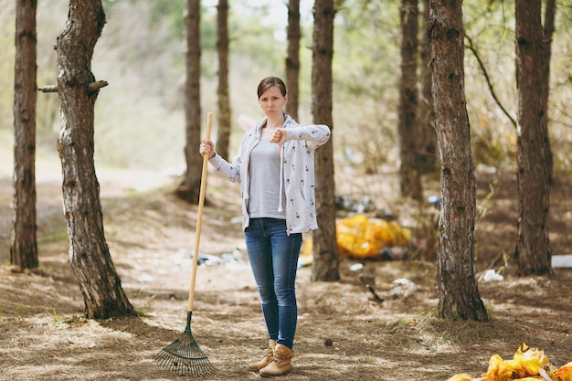 쓰레기 수거를 위해 갈퀴를 사용하여 쓰레기를 청소하는 젊은 화난 여성이 흩어져 있는 공원에서 엄지손가락을 아래로 보여줍니다. 환경 오염 문제. 자연 쓰레기, 환경 보호 개념을 중지합니다.