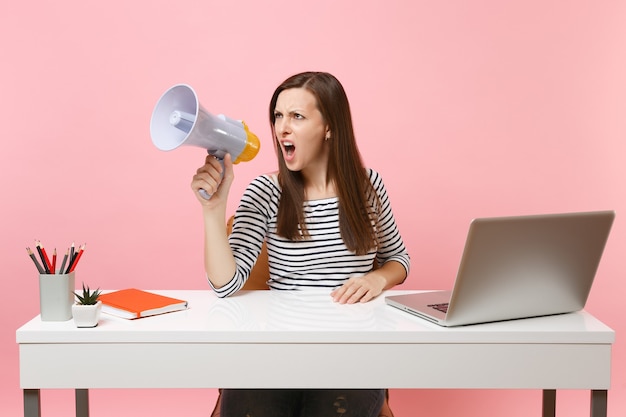 Young angry woman screaming in megaphone while sit, working on project at office with pc laptop