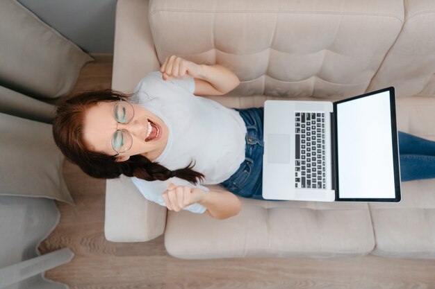 Молодая злая женщина в очках с помощью ноутбука дома в гостиной сидит на диване, работая ...